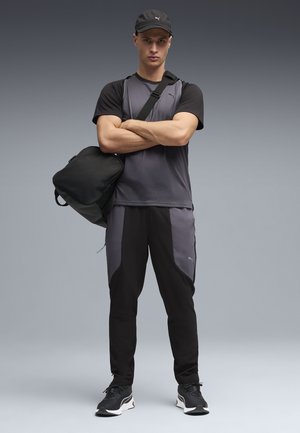 Sportliches Outfit mit einem grau-schwarzen Kurzarmshirt, passenden Hosen mit Akzenten und einer schwarzen Mütze. Eine schwarze Tasche hängt über der Schulter.