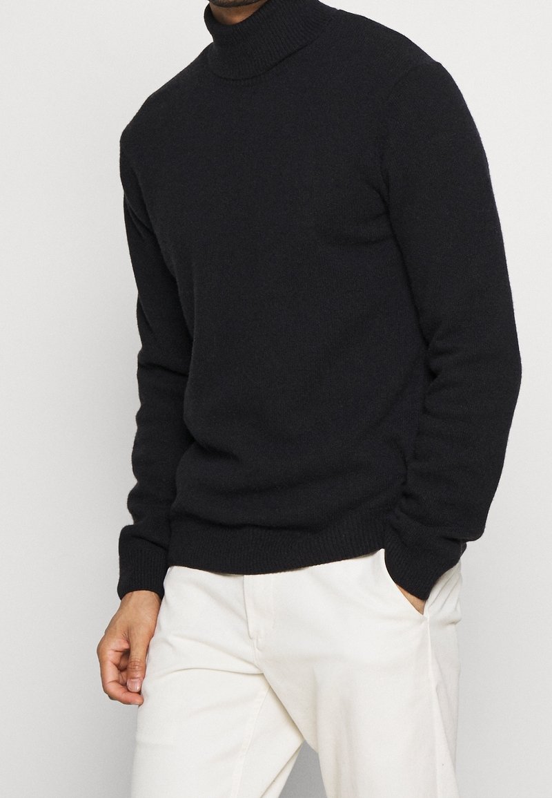 Maglione dolcevita nero, tessuto a maglia, design aderente, polsini e orlo a coste, indossato con pantaloni color crema. Nessun motivo o accento particolare.