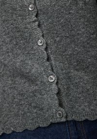 Pelēks filca kardigans ar viļņotu apakšu, tam ir četri pogas un mīksta tekstūra, daļēji ieliktas tumšā džinsu bikšu iekšpusē.