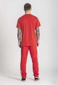 Muž s tetovaniami na oboch rukách, oblečený v červenom tričku a zodpovedajúcich červených nohaviciach s bielymi bočnými pruhmi, stojí otočený chrbtom.