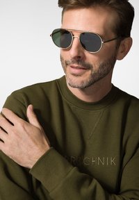 Zielony sweter z subtelną fakturą i wytłoczonym haftem. Projekt z okrągłym dekoltem, uzupełniony o okulary przeciwsłoneczne w złotej oprawie z ciemnymi szkłami.