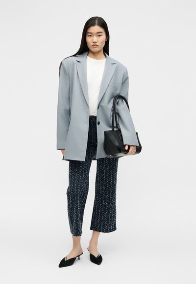 Femme portant un blazer gris clair oversize, chemise blanche, pantalon court bleu marine à motifs, talons noirs pointus, tenant un sac à main noir.