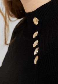 Pull en tricot noir avec un col montant et cinq boutons dorés texturés le long de l'épaule. Texture lisse avec des accents côtelés.