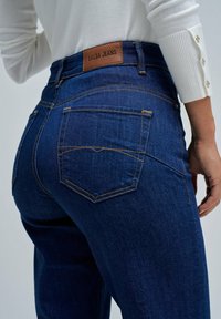 Mujer vistiendo unos Jeans Salsa de color azul oscuro con costuras naranjas y una camiseta blanca de manga larga, mostrada desde la cintura hacia abajo en un ángulo.
