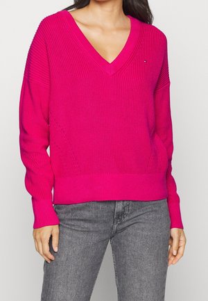 Pull en V rose avec un motif tricoté texturé, manches longues, poignets et ourlet côtelés. Associé à un jean en denim gris.