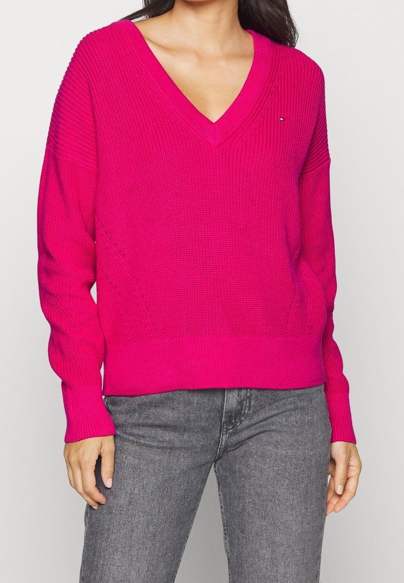 Rosa V-Ausschnitt-Pullover mit einem strukturierten Strickdesign, langen Ärmeln, gerippten Bündchen und Saum. Kombiniert mit grauen Jeans.