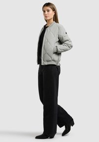 Femme debout les mains dans les poches, portant une veste matelassée gris clair, un haut noir, un pantalon large noir et des bottes noires sur un fond clair.