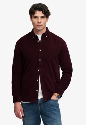 Mand med mørkt krøllet hår iført en mørkerød skjorte med knapper over en hvid t-shirt og blå jeans, med et ur på venstre håndled.