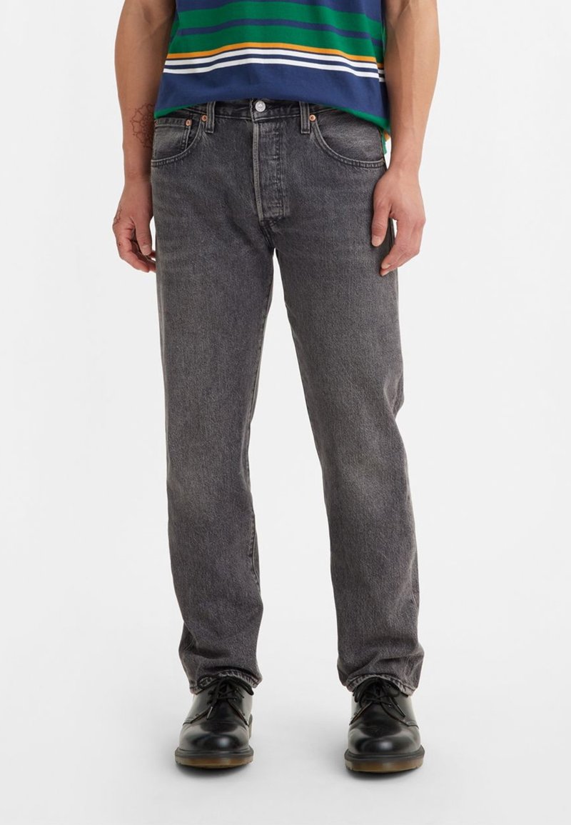 Personne portant un jean noir délavé à jambes droites, une chemise rayée vert, bleu, blanc et jaune, et des chaussures noires à lacets sur un fond blanc.