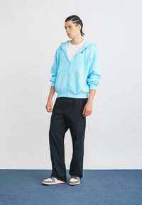 Φωτεινό μπλε φούτερ με φερμουάρ και μπροστινή τσέπη, διακοσμημένο με κεντημένο λογότυπο, συνδυασμένο με μαύρα παντελόνια και γκρι αθλητικά παπούτσια με πολύχρωμα κορδόνια.