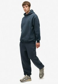 Mørkeblå hættetrøje med kenguru-lomme og ribbede manchetter, parret med matchende tapered sweatpants. Stoffet ser blødt, børstet og behageligt ud.