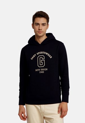 Young man wearing a black GANT Sportswear hoodie with "New Haven 1949" text, standing against a plain white background.