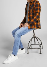 Person som sitter på en metallpall, iklädd ljusblå jeans, vita sneakers, grå t-shirt och en skjorta i orange och marinblå rutmönster.