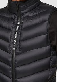 Sort puffervest med høj krave, lynlåsgreb foran og vandret quiltning. Har kontrasterende tekst langs lynlåsen. Glat og let tekstur.