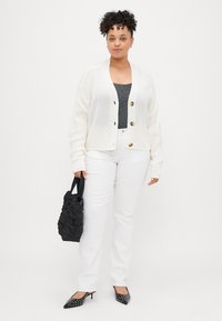 Frau steht da, trägt eine weiße Strickjacke, ein dunkles Oberteil, weiße Hosen, schwarze, mit Nieten verzierte Absätze und hält eine schwarze, geraffte Handtasche vor einem weißen Hintergrund.