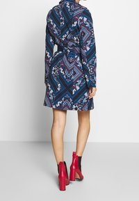 Niebieska sukienka w wzory z długimi rękawami, ozdobiona skomplikowanymi wzorami w bieli, różu i purpurze. Połączona z czerwonymi botkami na obcasie.