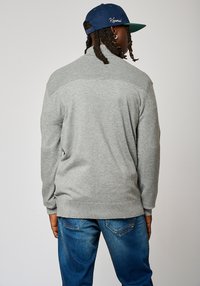 Suéter de punto gris con un collar alto, detalles acanalados en los hombros y mangas alargadas, combinado con jeans de mezclilla azules.