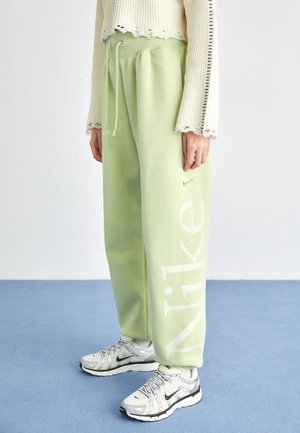 Personne portant un pantalon de survêtement Nike vert clair avec un grand logo blanc et des baskets blanches avec des détails noirs sur un sol bleu.