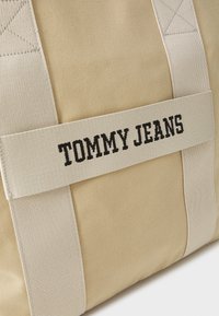 Bež torba od tkanine s kremastom trakom koja ima crni izvezeni natpis "TOMMY JEANS." Vidljive šavove i teksturiranu površinu.