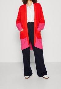 Osoba ubrana w białą koszulę na guziki, czerwono-różowy długi kardigan, granatowe spodnie o szerokich nogawkach oraz białe trampki na jednolitym tle.