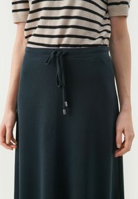 Jupe bleu marine avec une taille à cordon de serrage, dotée de pointes en métal argenté à ses extrémités. Assortie à un haut en tricot rayé beige et noir.