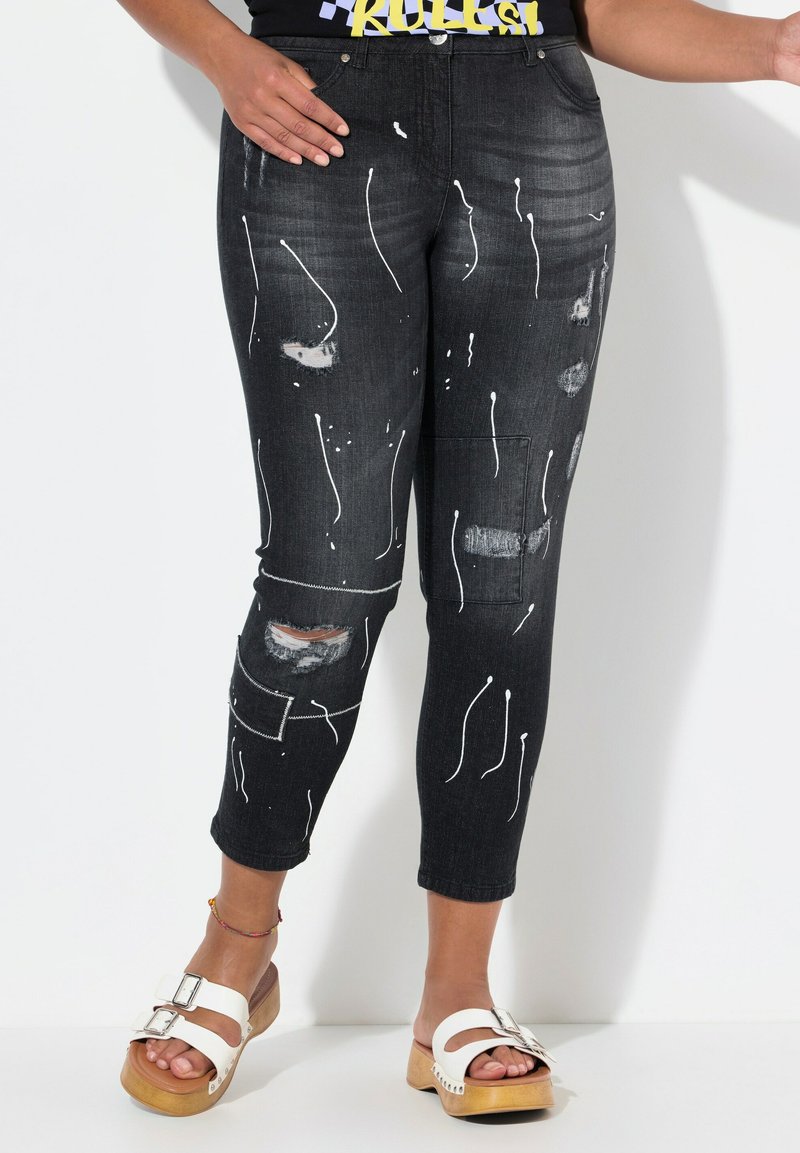 Vaqueros de mezclilla negra con salpicaduras de pintura blanca y detalles en desgaste, que presentan un ajuste slim y un largo recortado. Costuras blancas y bolsillos.