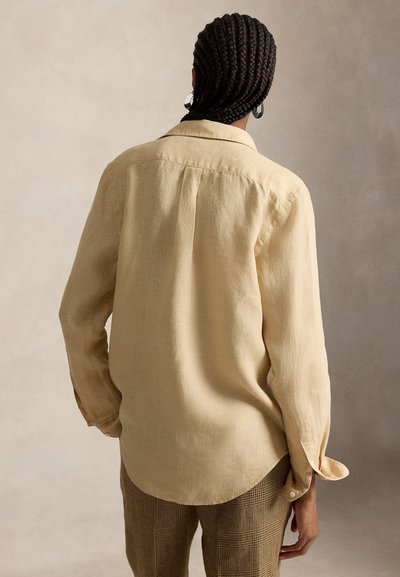 Femme aux cheveux tressés portant une chemise beige à manches longues et un pantalon à carreaux, debout de dos à la caméra sur un fond neutre.