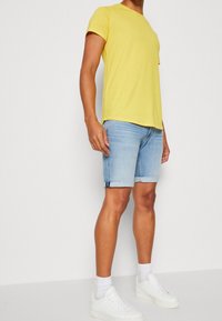 Camiseta amarilla de manga corta combinada con pantalones cortos de denim azul claro con dobladillos enrollados. Zapatillas blancas y calcetines tobilleros completan el atuendo.