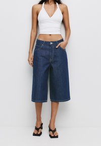 Pantalon culotte en denim foncé à coupe décontractée, doté de poches avant et d'un ourlet cousu. Associé à un crop top blanc et des sandales noires.