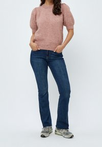 Žena v růžovém svetru s krátkým rukávem, modrých rovně střižených džínech a šedých teniskách, stojící s rukama v kapsách na bílém pozadí.