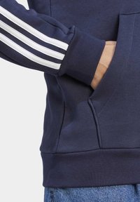 Laivastonsininen huppari, jossa valkoinen kolmiraidallinen kuvio hihoissa, resorit hihansuuissa ja etutasku; valmistettu pehmeästä, teksturoidusta materiaalista.