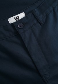 Navy blue trousers with a smooth texture, featuring a button closure and a stitched label with a logo. Detailed seams at the waistband.