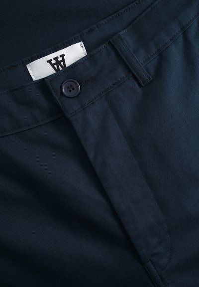 Pantalons bleu marine avec une texture lisse, comprenant une fermeture à bouton et une étiquette cousue avec un logo. Coutures détaillées à la taille.