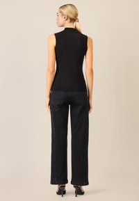 Donna con coda di cavallo bionda, che indossa un top senza maniche nero, jeans neri a gamba larga con borchie e sandali neri con tacco alto, in piedi su uno sfondo beige.