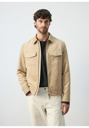 Hombre de pie con chaqueta beige con cremallera y bolsillos con solapa, camisa negra y pantalones blanco sucio contra un fondo claro y liso.