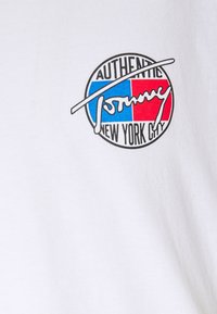 Bílá látka s kulatým logem obsahujícím modré a červené bloky, slova "AUTHENTIC," "NEW YORK CITY" a stylizovaný nápis "Tommy".