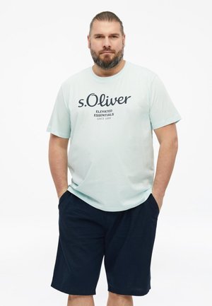Mężczyzna z brodą i związanymi włosami, ubrany w jasnoniebieską koszulkę "s.Oliver Elevated Essentials" oraz ciemnoniebieskie spodenki, ręce w kieszeniach, neutralne tło.