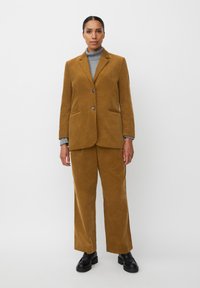 Blazer in velluto a coste senape abbinato a pantaloni. Il blazer presenta due bottoni e tasche; sotto c'è un dolcevita grigio. Scarpe nere completano l'insieme.
