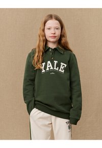 Grønn bomullssweatshirt med "YALE"-logo i hvitt, knappet krage og ribbestrikkede mansjetter. Brukt med lyse bukser med logo-detalj.