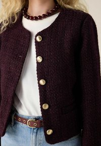 Bordeaux kleurige gestructureerde jasje met gouden knopen, ronde halslijn en een klein vakje. Gecombineerd met een witte blouse en een bruine riem over denim.