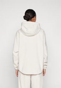 Vrouw gezien van achteren in een witte fleece hoodie en bijpassende witte broek tegen een effen witte achtergrond.