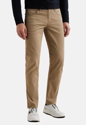 Mężczyzna w beżowych, dopasowanych spodniach i białych trampkach, stojący z rękami swobodnie opuszczonymi wzdłuż ciała na jednolitym tle.