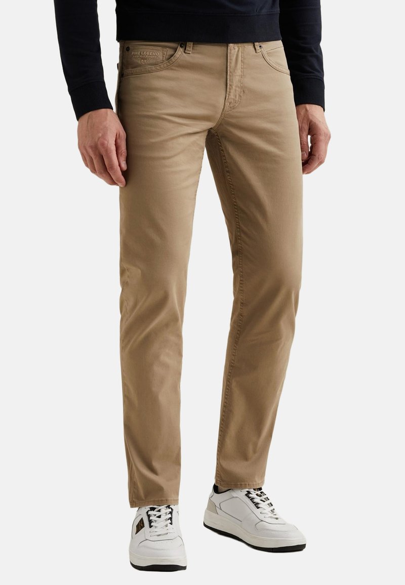 Mann trägt beige, eng anliegende Hose und weiße Turnschuhe, steht mit entspannten Händen seitlich an einem einfarbigen Hintergrund.