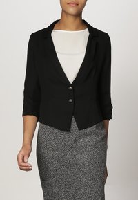 Blazer noir cintré avec des manches trois-quarts, deux boutons ronds et une coupe ajustée, associé à une jupe grise texturée.