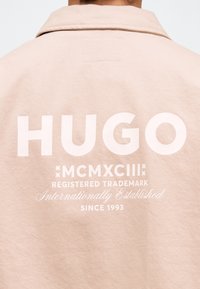 Άτομο που φοράει μπεζ μπουφάν με μεγάλο λευκό κείμενο "HUGO" και μικρότερο κείμενο "MCMXCIII Registered Trademark Internationally Established Since 1993" στην πλάτη.