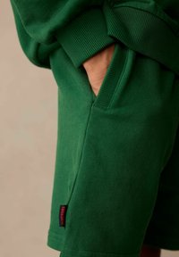 Gröna bomullsshorts med en ribbad midja, sidofickor och en liten röd "Superdry"-etikett på fållen. Slät yta, sportig design.