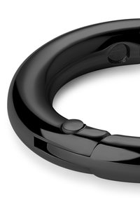 Svart sirkulært objekt med glatt, blank overflate. Har en knapp og en spalte på den ene siden, med et stilrent, minimalistisk design.