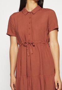 Robe couleur rouille avec un col, des manches courtes, une fermeture à boutons à l'avant et une ceinture à la taille ; présente une jupe à volants avec une texture lisse.