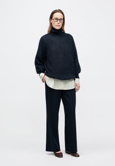 Mørkeblå sweater med høj krave og ballonærmer, kombineret med en lysegrøn skjorte med krave og marineblå bukser med nålestriber. Bourgognefarvede flade sko.