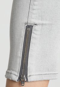 Pantalones cortos de mezclilla gris claro con una cremallera lateral que tiene un tirador plateado YKK. La textura de la tela es suave, con un diseño ajustado y puños doblados.